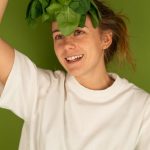 Cheerful adult female in white t shirt with fresh spinach foliage against forehead looking up